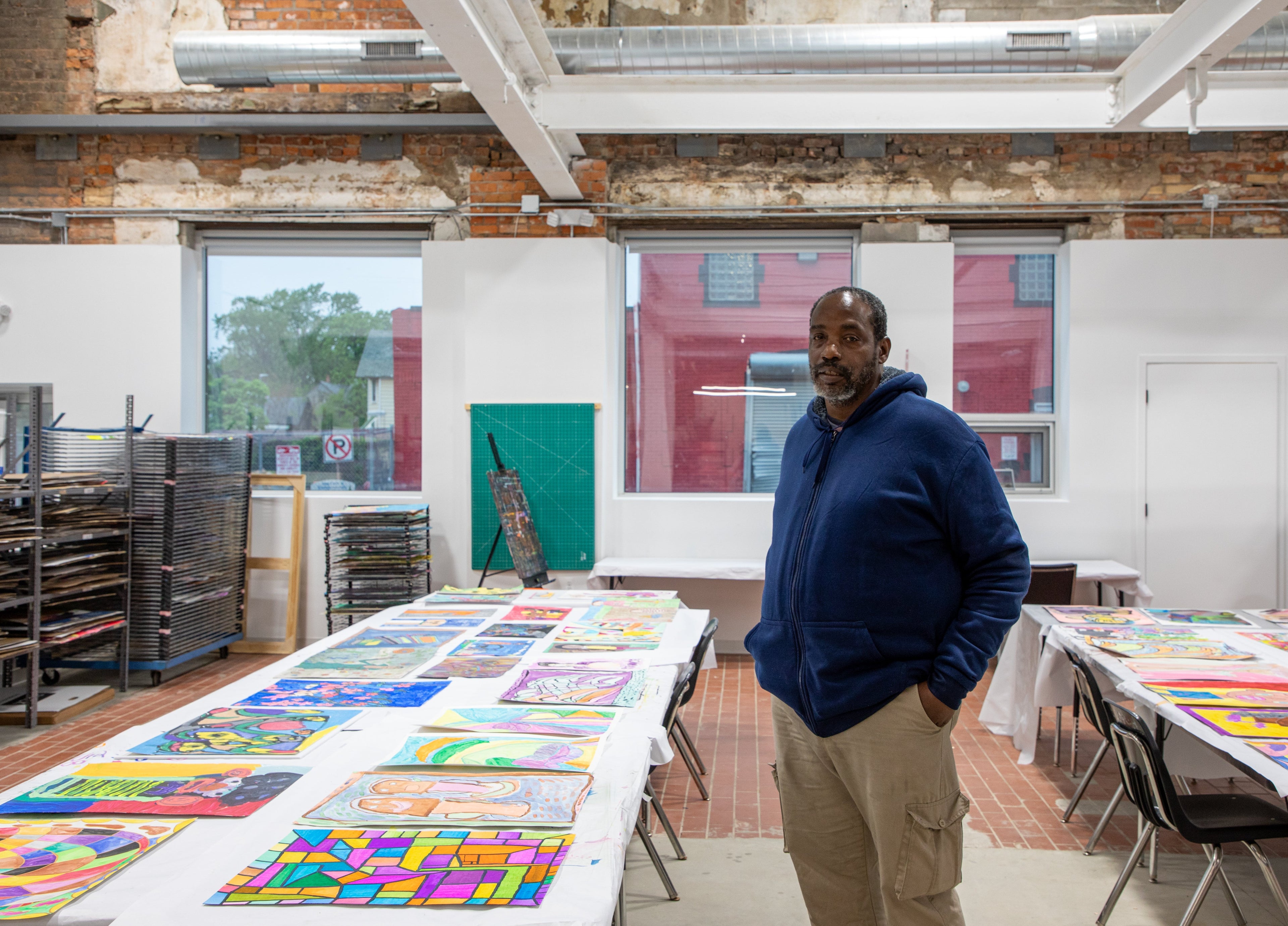 Ronald Griggs stands in "If Anyone Can Hear This" exhibit, wearing a dark blue hoodie. Behind him are two framed pieces: a figure sketch with blue background above, and a more detailed, surreal painting of human forms below.