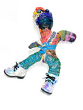 A colorful, paint-splattered doll figure posed dynamically on a plain white background. The figure has a multicolored body with blue, purple, red, green, yellow, and orange paint textures, white shoes with iridescent blue and pink laces, and black shoes on one foot.